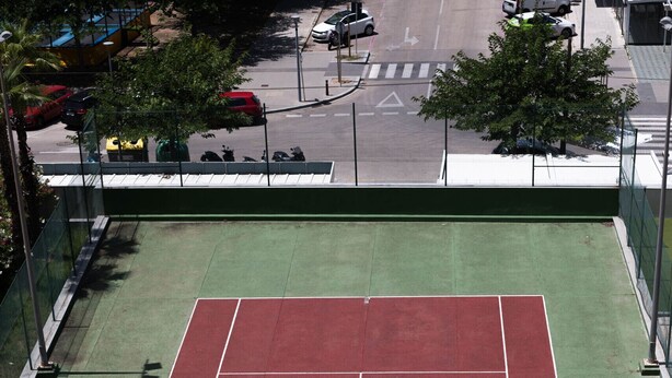Uma quadra de tênis com rede, cercada por uma grade verde e uma calçada.