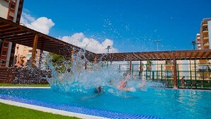 Piscina com pessoas brincando, rodeada por edifícios e um céu claro.