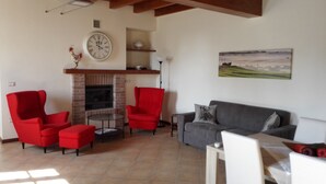 Un salon avec une cheminée, des fauteuils rouges, un canapé gris et un tableau au mur.