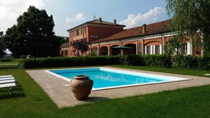 Une piscine avec un grand pot en terre cuite au premier plan, un bâtiment doté de fenêtres en arc de cercle et d’une façade rouge, et une pelouse bien entretenue.