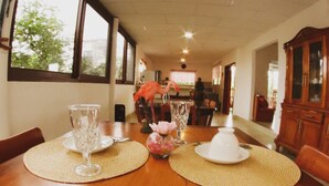 A dining area with a wooden table set for two, a vase with pink flowers, and a view of the kitchen in the background. - Casa Hospedaje Punta Arena (Puerto Villamil)