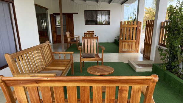A wooden bench with a cushioned seat and backrest, a wooden chair with a cushion, and a small round table in a room with green artificial turf flooring. - Casa Hospedaje Punta Arena (Puerto Villamil)
