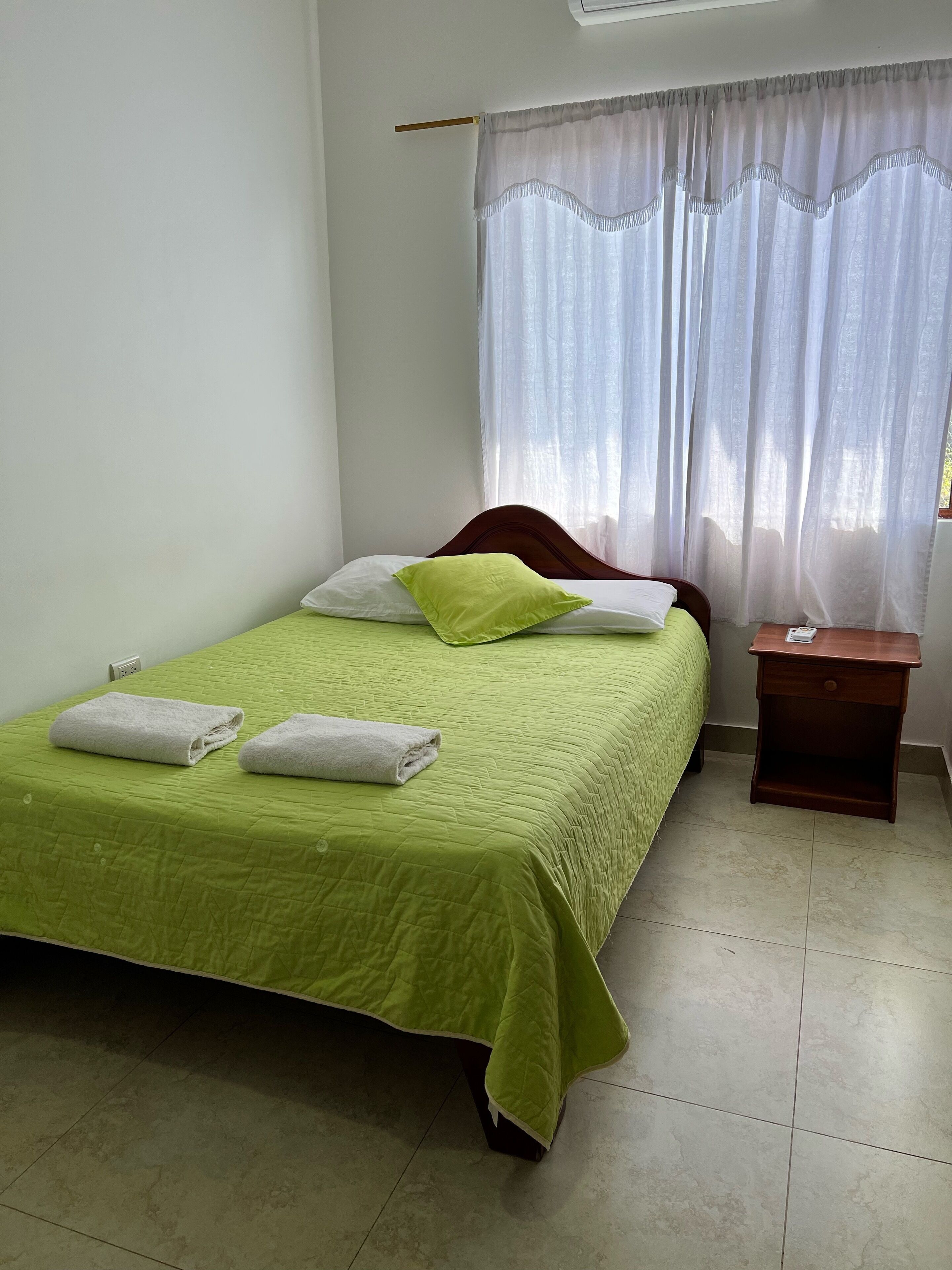 A neatly made bed with a green bedspread and two white towels, a wooden nightstand with a book, and a window with sheer curtains.