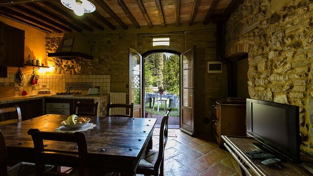 A rustic kitchen with a stone wall, wooden beams, a dining table with chairs, a television, and a view through an open doorway. - Apartment/ flat - Chianni (Chianni)