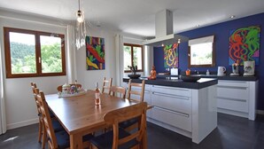 Una cocina moderna con una isla central, una mesa de comedor de madera y sillas. Hay pinturas abstractas coloridas en las paredes, y se ve vegetación a través de las ventanas.