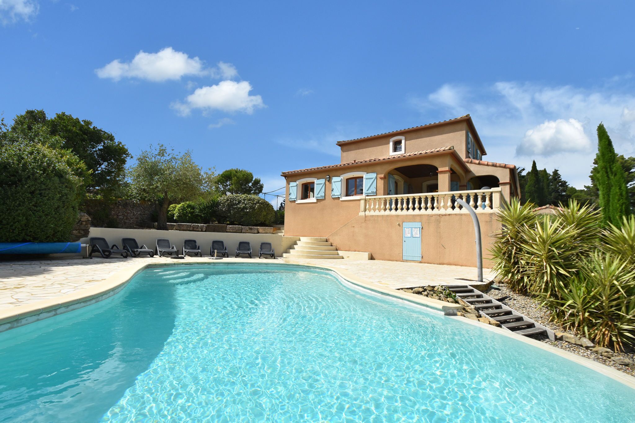 A swimming pool with lounge chairs, a building with a balcony, and a clear blue sky.
