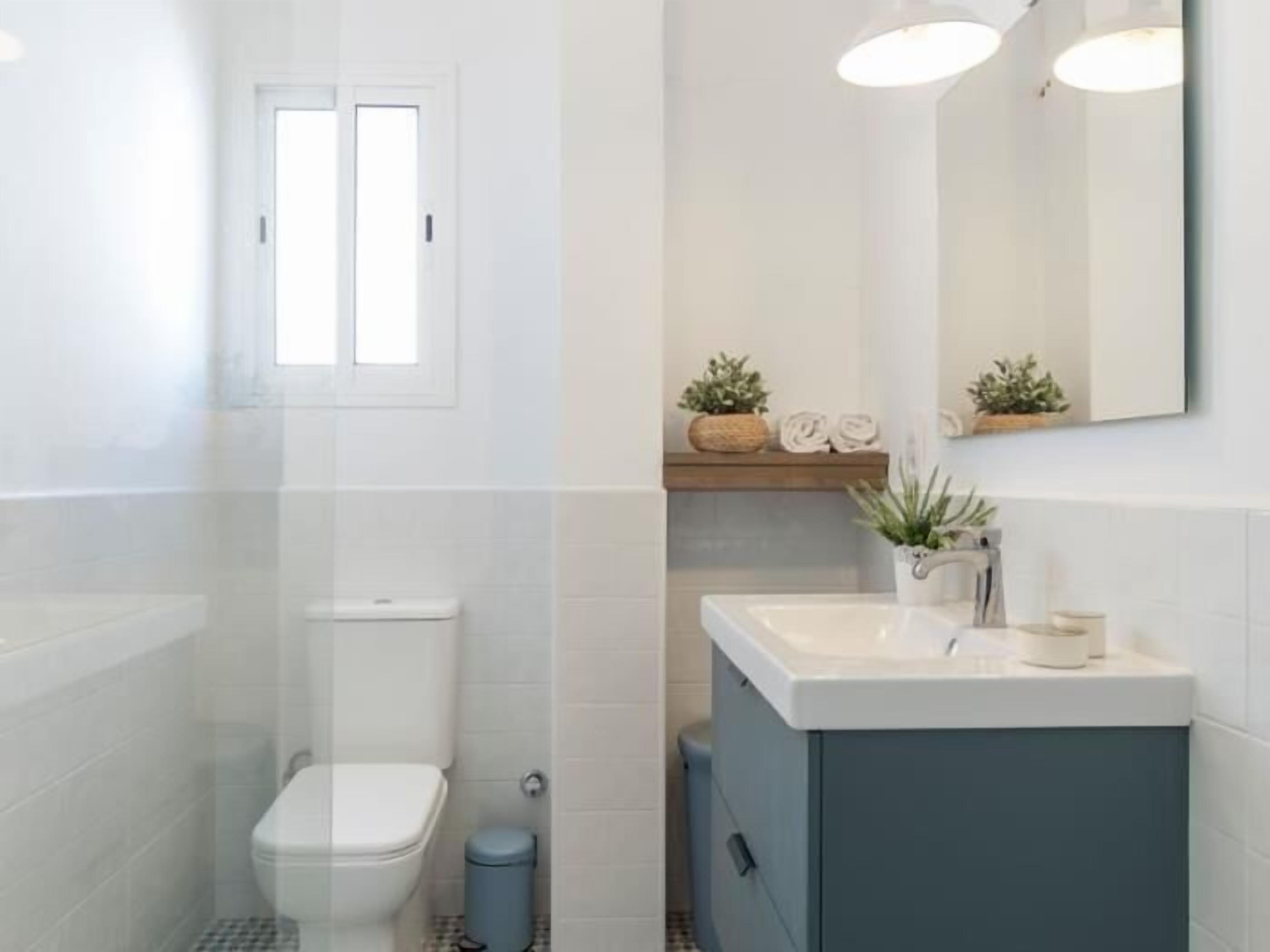 A modern bathroom with a toilet, sink, and cabinet. There is a window above the toilet and a mirror above the sink.