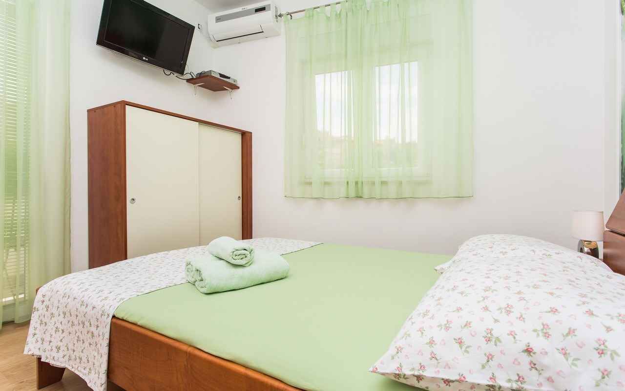 A bedroom with a bed, a wardrobe, a television, and a window with sheer curtains.