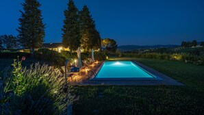 A rectangular swimming pool with a wooden deck and lounge chairs, surrounded by tall trees and a well-lit garden area. - Exclusive Estate | Personal Concierge & Heated Pool (impruneta)