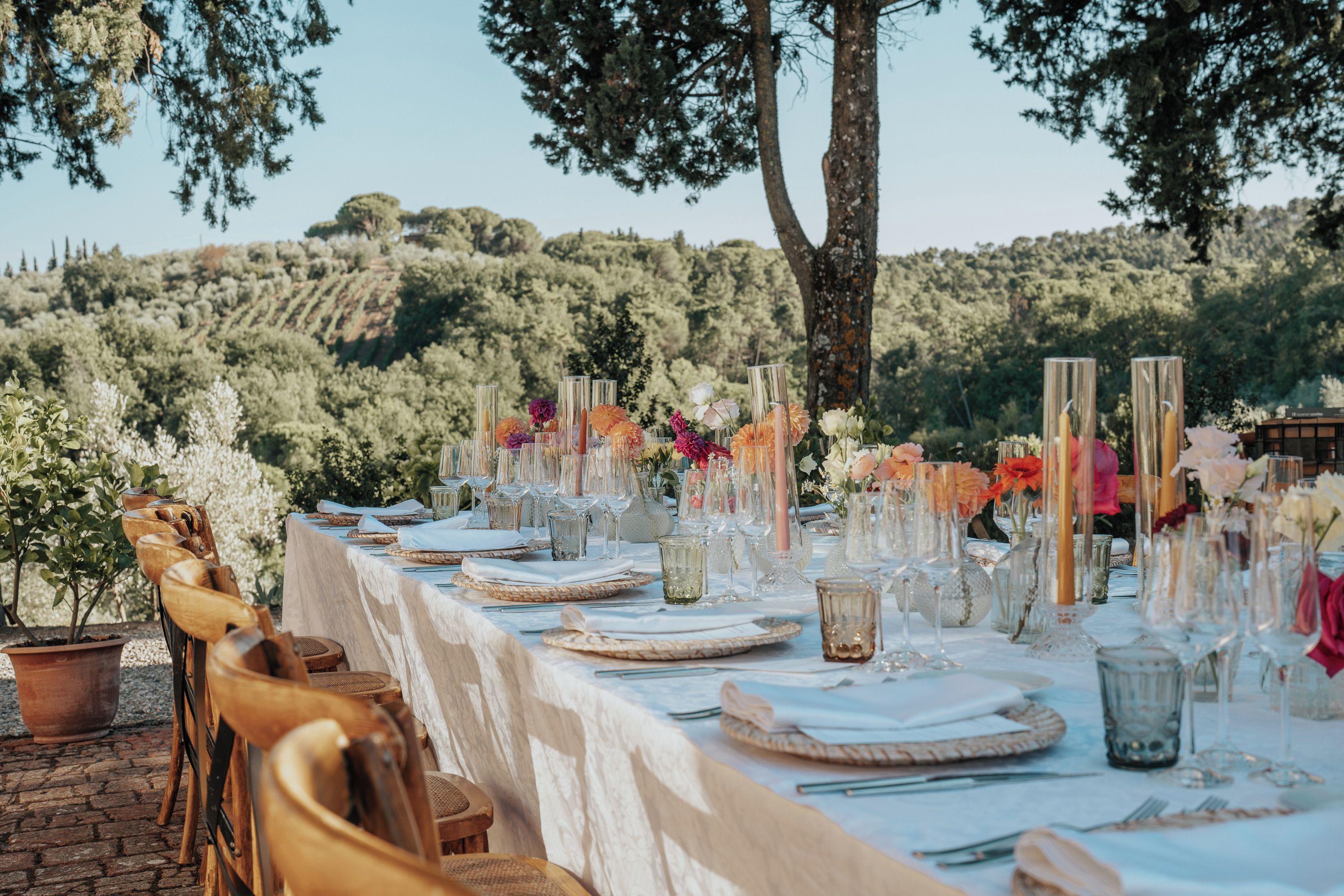 An outdoor dining area with a long table set for a meal, surrounded by trees and hills.