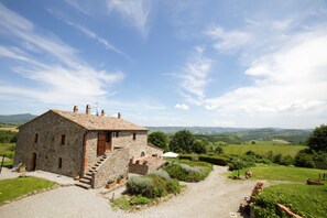 A stone building with a tiled roof, a gravel driveway, and a view of rolling hills in the background. - Large double bedroom: private bathroom, private access, garden and swimming pool (Radicofani)