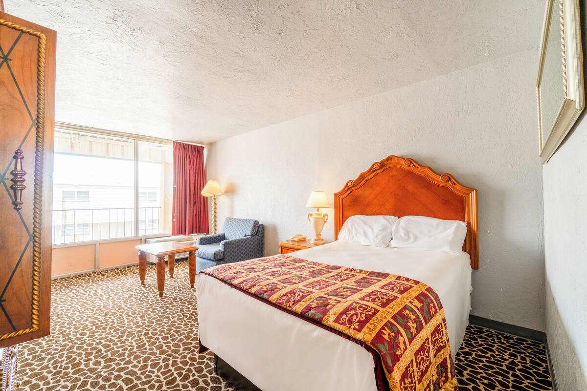 Una habitación de hotel con una cama grande, una mesita pequeña, una silla, un ventanal y una puerta.