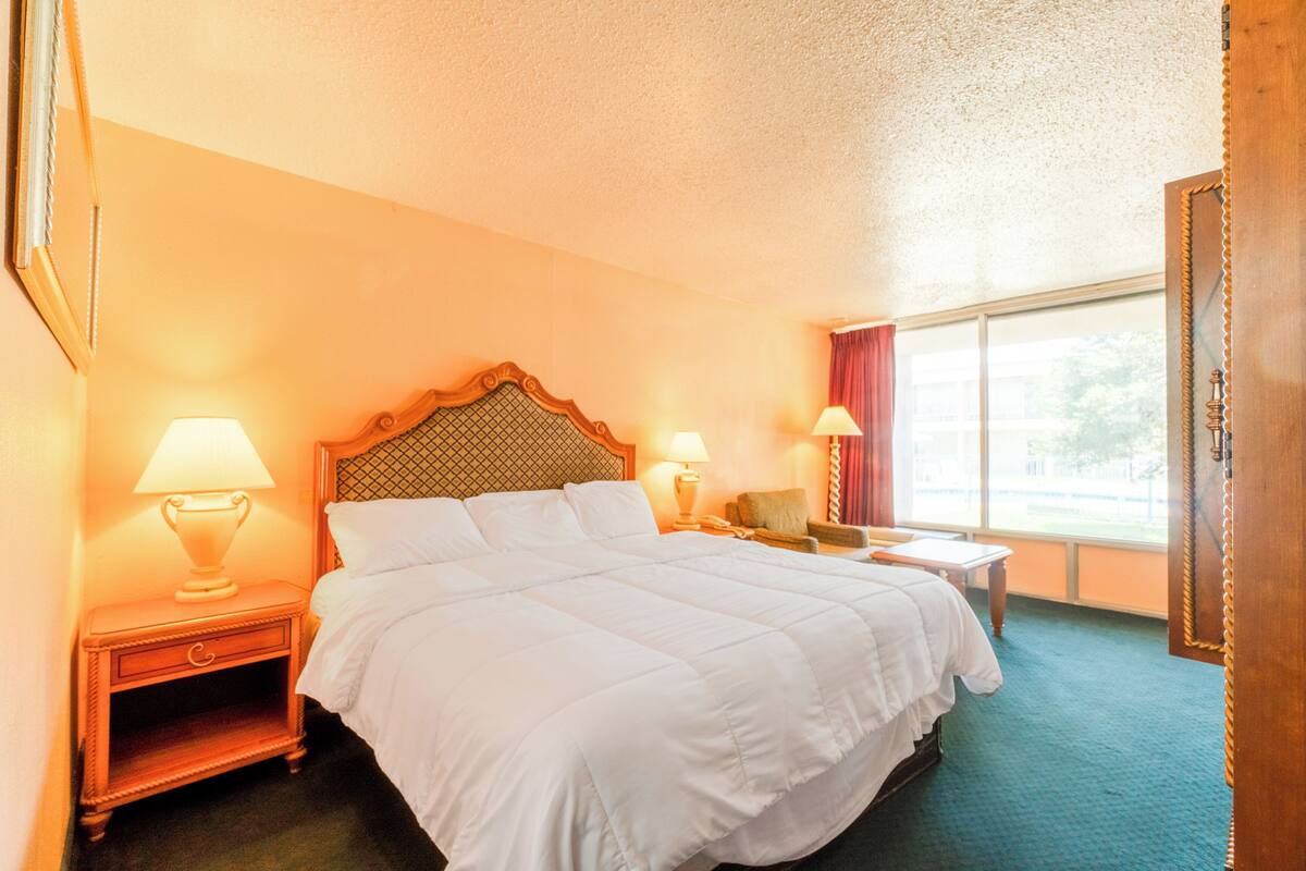 Habitación de hotel con una cama grande, dos mesitas de noche con lámparas, una mesita auxiliar, una ventana con vistas y una puerta de madera.