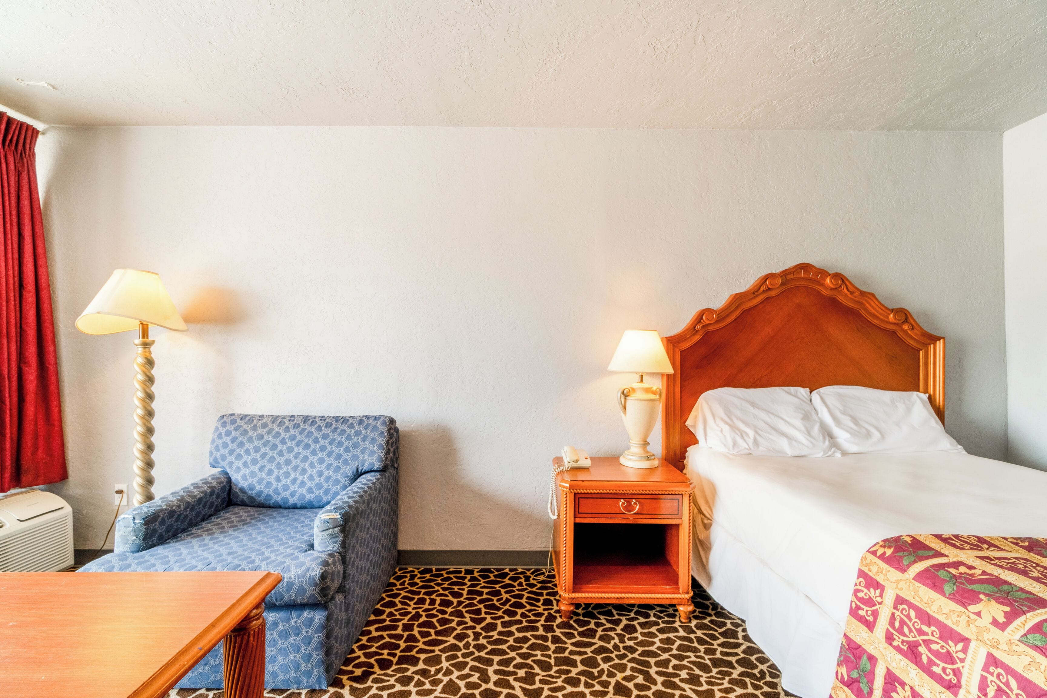 A hotel room with a bed, a chair, a nightstand with a lamp, and a patterned carpet.