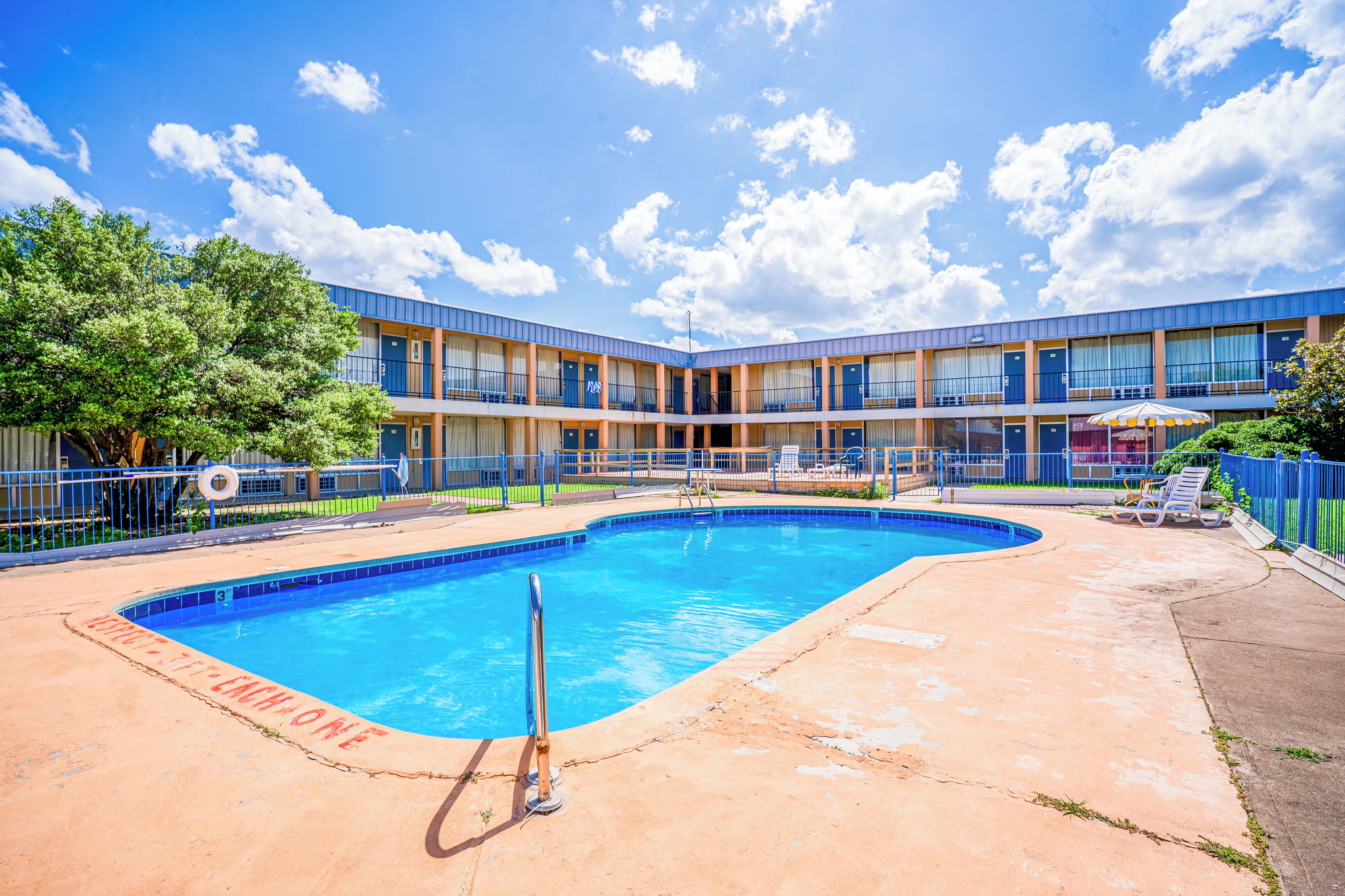 A swimming pool with a blue border and a lifeguard stand, surrounded by a building with multiple balconies and a fenced area with lounge chairs.