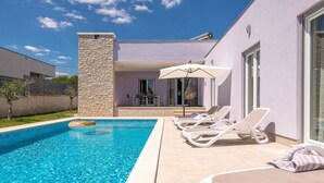 Uima-allas, jossa on aurinkotuoleja ja aurinkovarjo, kivipilari sekä terassialue, jossa on pöytä ja tuolit.