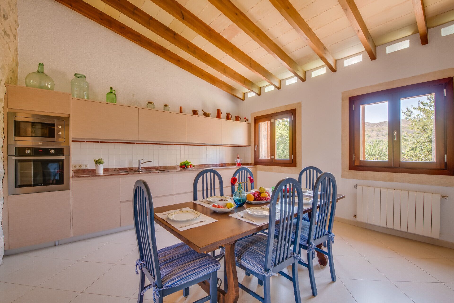 A kitchen with wooden beams, a dining table with chairs, and a window with a view of greenery.