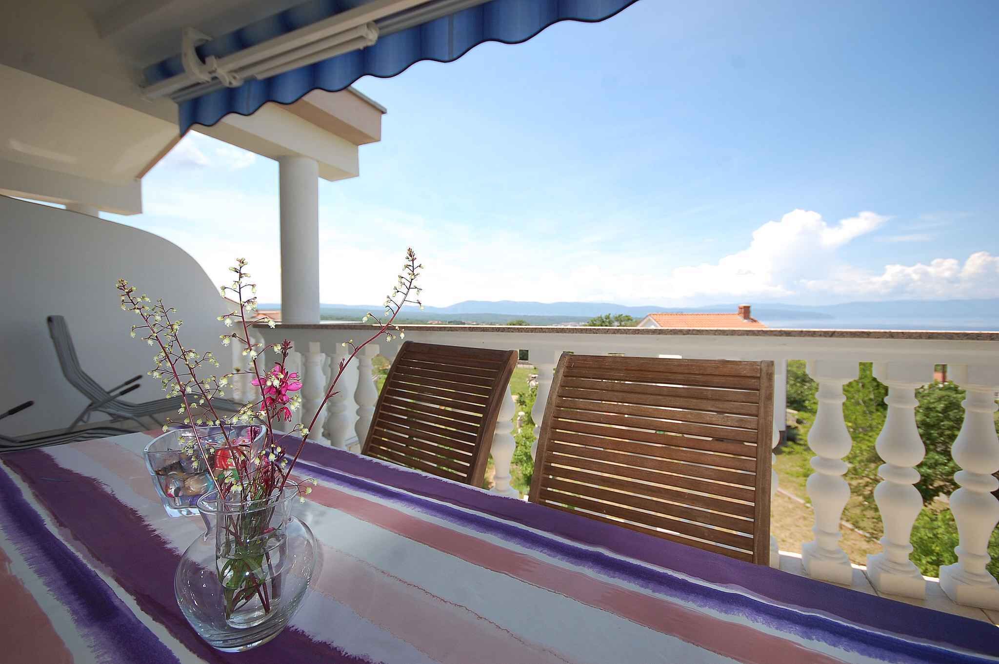 Un balcón con vistas a montañas distantes, un florero con flores y sillas de madera.