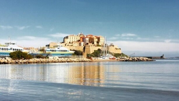 A coastal building with a stone facade, a clear sky, and calm water reflecting the structure. - Studio Orsoni - See View (Calvi)