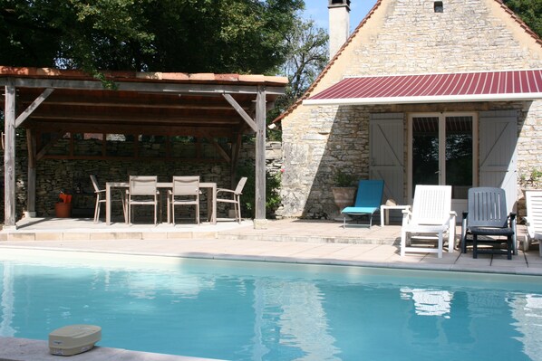 Un bâtiment en pierre avec un toit rouge, un patio couvert avec des chaises et une piscine.
