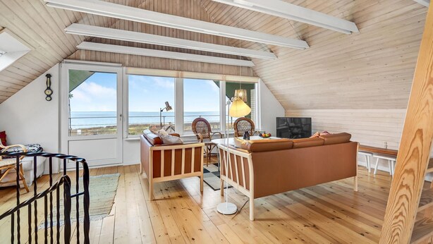 Un acogedor cuarto en ático con techo de madera, ventanas amplias con vistas al mar, un sofá marrón, una mesa de comedor y un televisor.