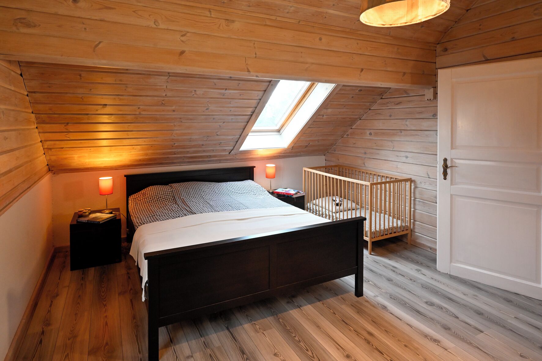 Une chambre avec un plafond en pente, un puits de lumière, un lit, une table de chevet avec une lampe, un berceau et une porte.