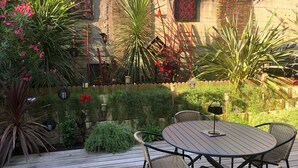 A wooden deck with a round table and four wicker chairs, surrounded by various plants and a wooden fence.