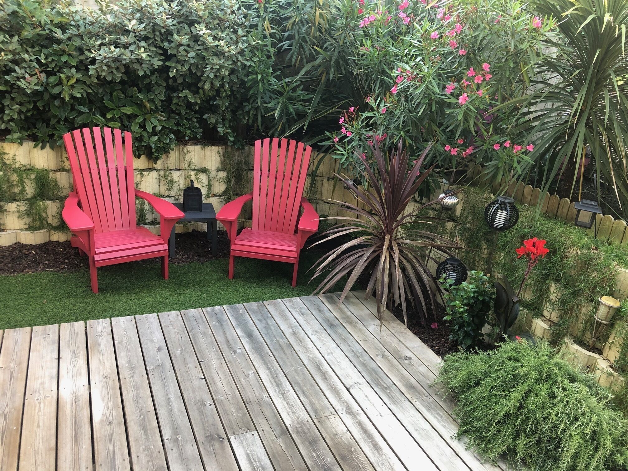 Eine Holzterrasse mit zwei roten Stühlen und einem kleinen Tisch, umgeben von üppigem Grün und verschiedenen Pflanzen.