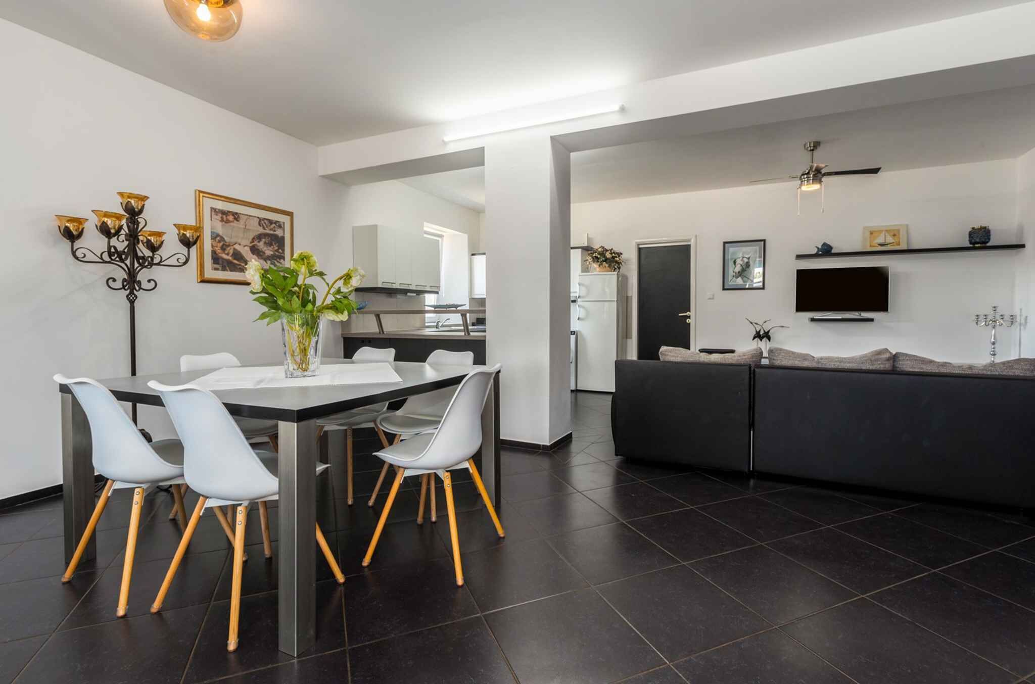 Eine moderne Küche mit dunklen Fliesen, ein Essbereich mit weißem Tisch und Stühlen sowie eine Wohnbereich mit Sofa und Fernseher.