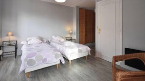 A bedroom with two beds, a wooden wardrobe, a white door, and a wicker chair.