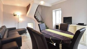 A modern living room with a dining area, featuring a staircase, a flat-screen TV, and a sofa.