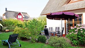 A patio with a table and chairs, a thatched roof, and a garden with bushes and flowers.