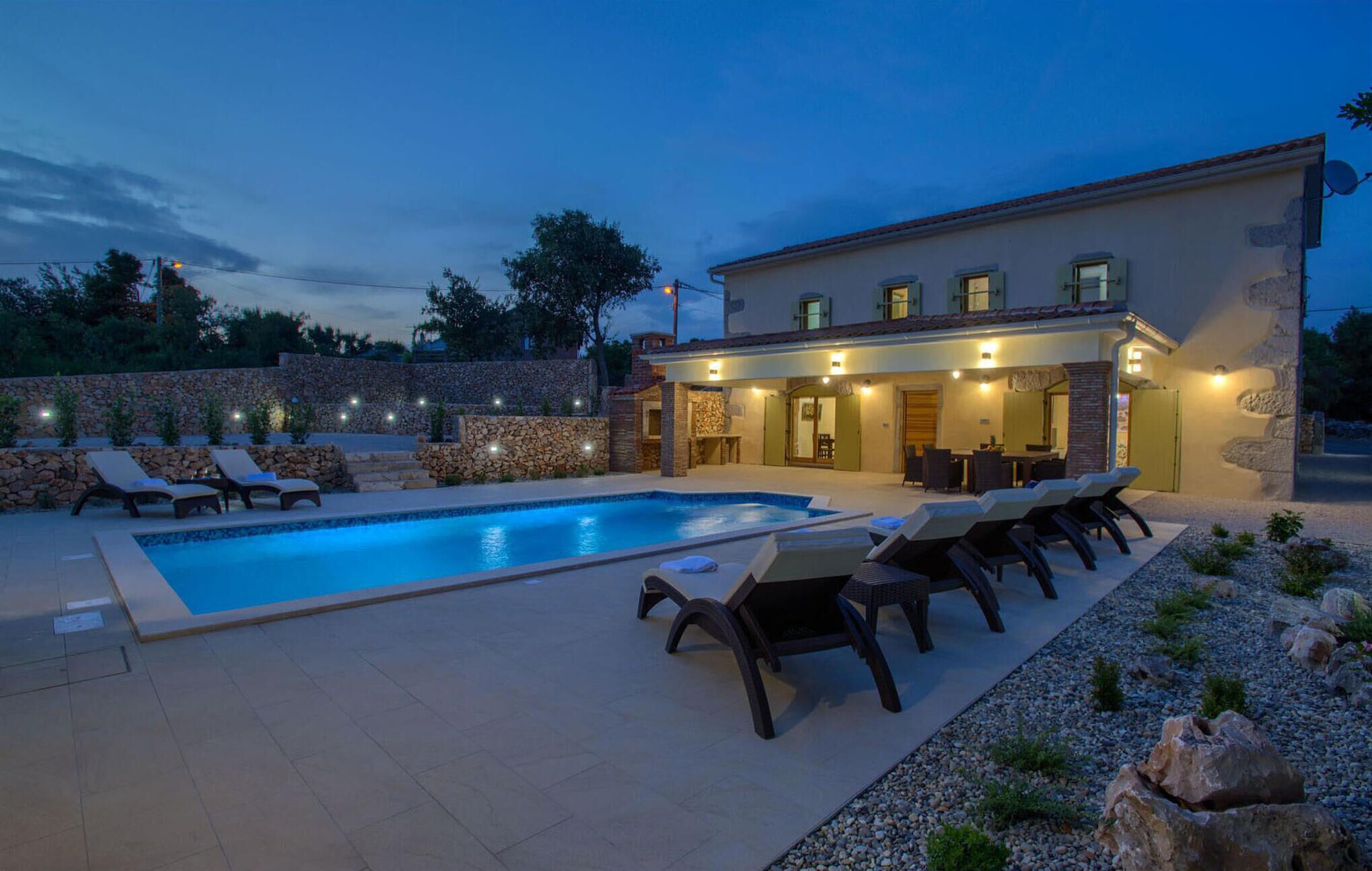 Une piscine avec des chaises longues, un mur en pierre et un bâtiment doté d’un éclairage extérieur.
