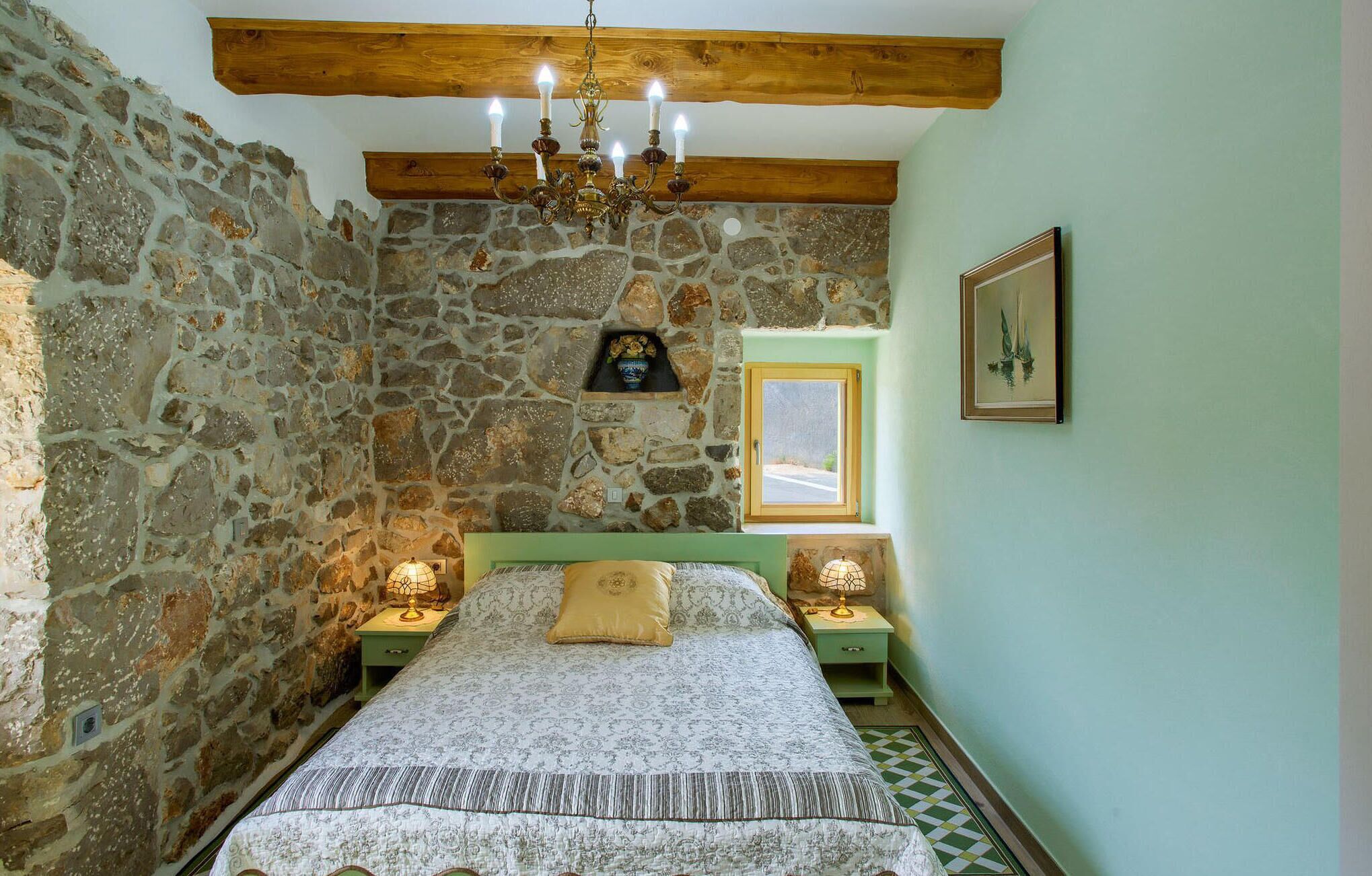 Een slaapkamer met een stenen muur, houten plafond, een bed met beddengoed met een patroon, twee nachtkastjes met lampen, en een lijst met een afbeelding aan de muur.