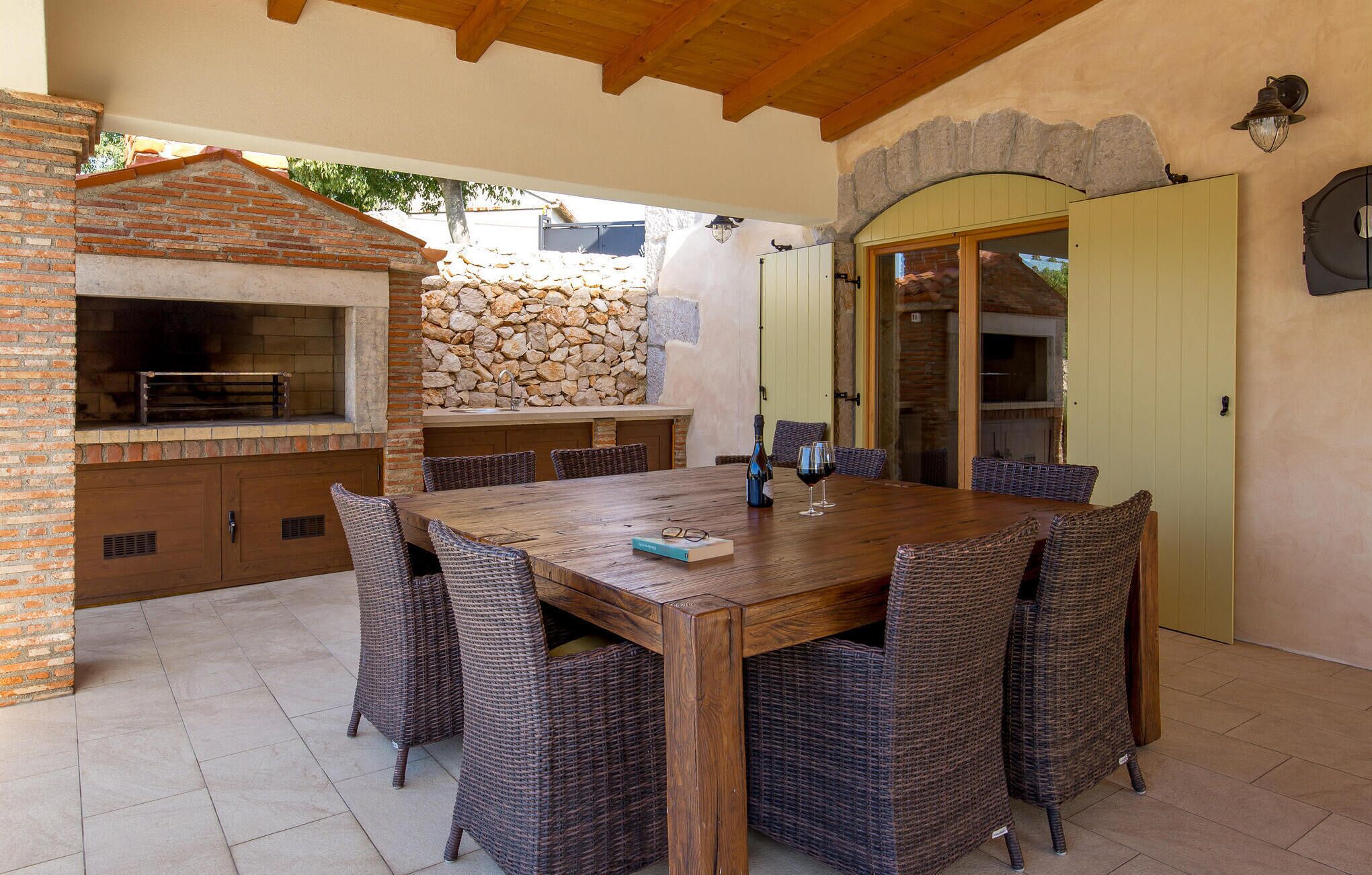 Een terras buiten met een bakoven, een stenen muur, rieten stoelen en een houten tafel.