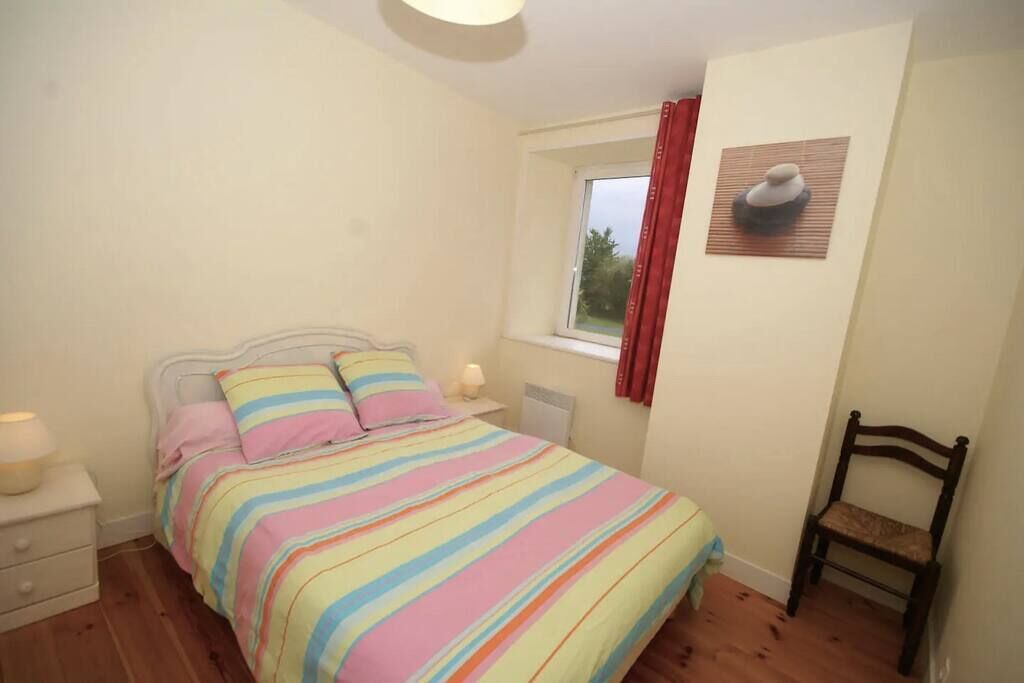 A bedroom with a bed, a nightstand, a chair, a window with curtains, and a wall-mounted picture.