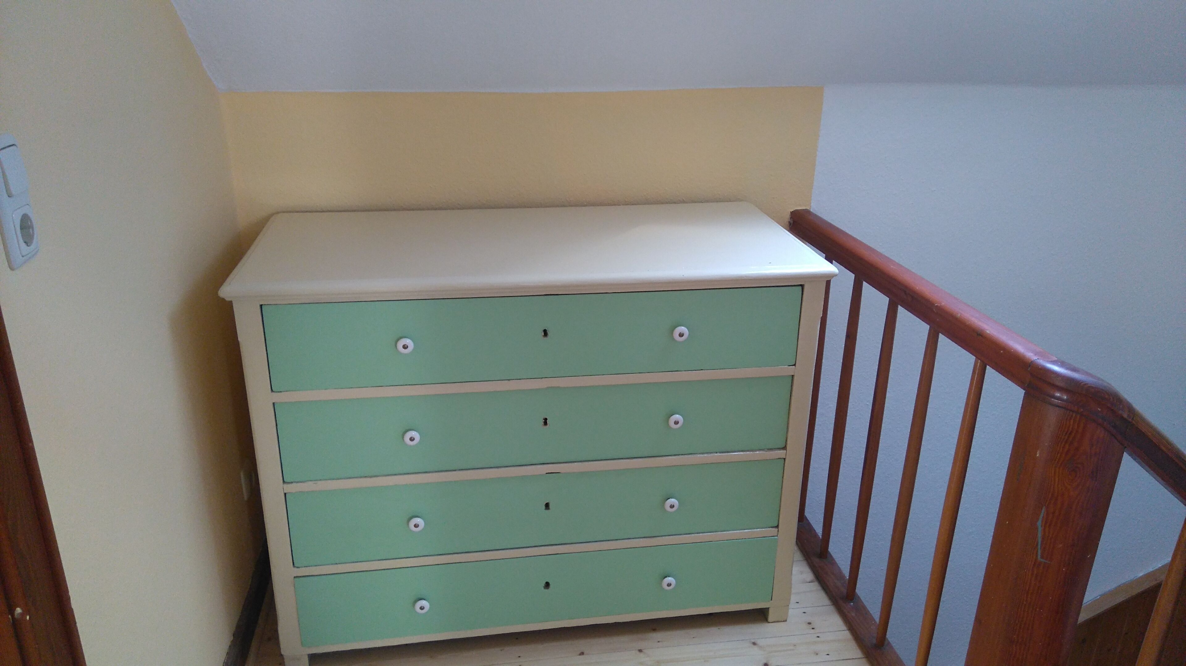 A green dresser with white drawers against a beige wall, located in a corner with a wooden railing.