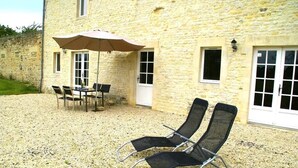 Un bâtiment en pierre doté d’un patio comprenant une table avec des chaises et un parasol, ainsi que deux fauteuils de jardin.