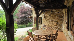 Een overdekt terras met een stenen muur, houten tafel en stoelen, en uitzicht op een tuin met bomen.