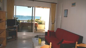 A room with a red sofa, a wooden chair, a small table, and a television on a stand. There is a sliding glass door leading to a balcony with a view of the sea. - 3 room apartment La Rochelle Les Minimes - Seaside (La Rochelle)