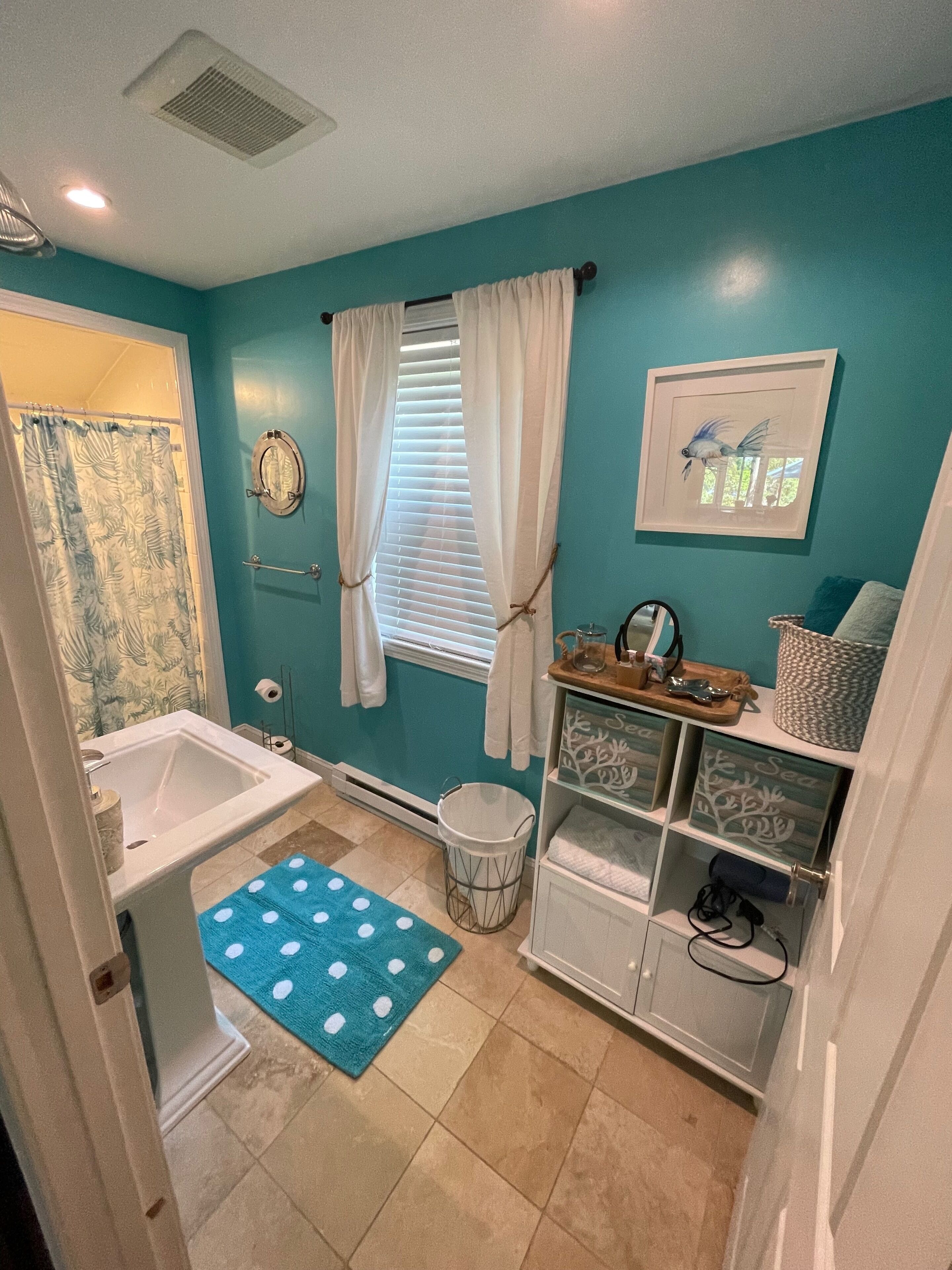 A bathroom with a bathtub, a toilet, a sink, a window with curtains, a towel rack, a framed picture, and a shelf with decorative items.