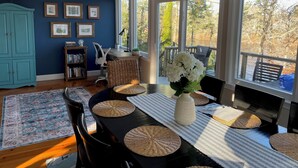A dining area with a table set for six, a blue wall with framed pictures, a teal cabinet, and a view of trees through large windows. - Enjoy the lake, dock, beach, large grounds, or renovated home.  New ductless AC. (East Falmouth)