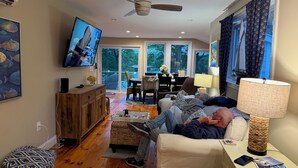A living room with a TV, wooden furniture, a ceiling fan, and a view through large windows. - Enjoy the lake, dock, beach, large grounds, or renovated home.  New ductless AC. (East Falmouth)