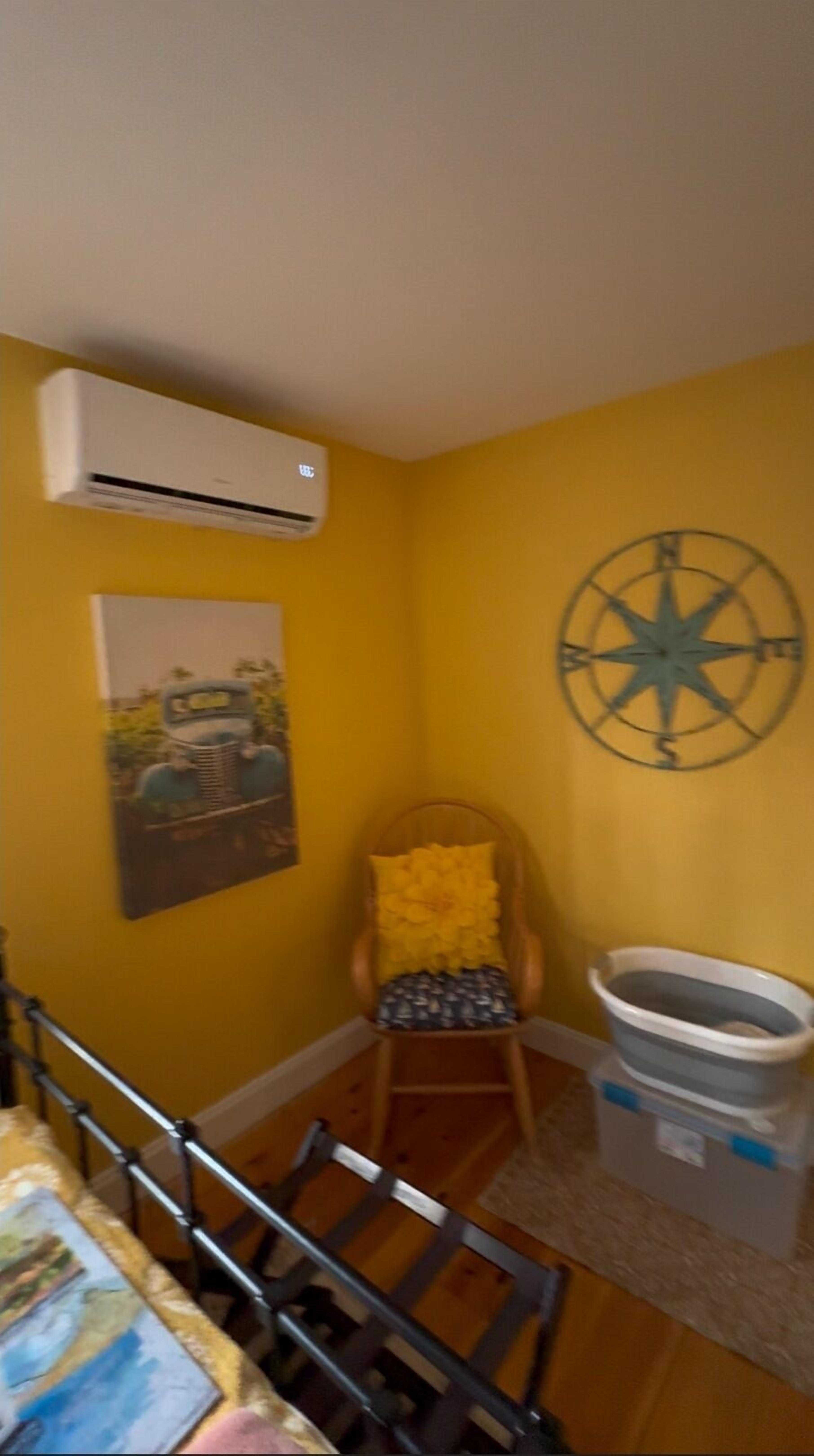 Une pièce avec un mur jaune, une chaise en bois avec un coussin jaune, un appareil de climatisation blanc, un objet décoratif en forme de boussole et un tableau.