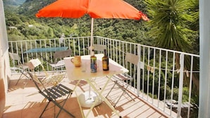 Ein Balkon mit Tisch und Stühlen, einem orangefarbenen Sonnenschirm und Blick auf eine gebirgige Landschaft.