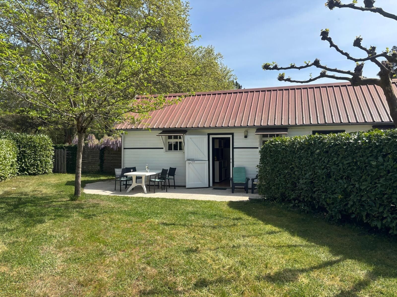A single-story building with a red roof, a small patio with a table and chairs, and a hedge in the foreground.
