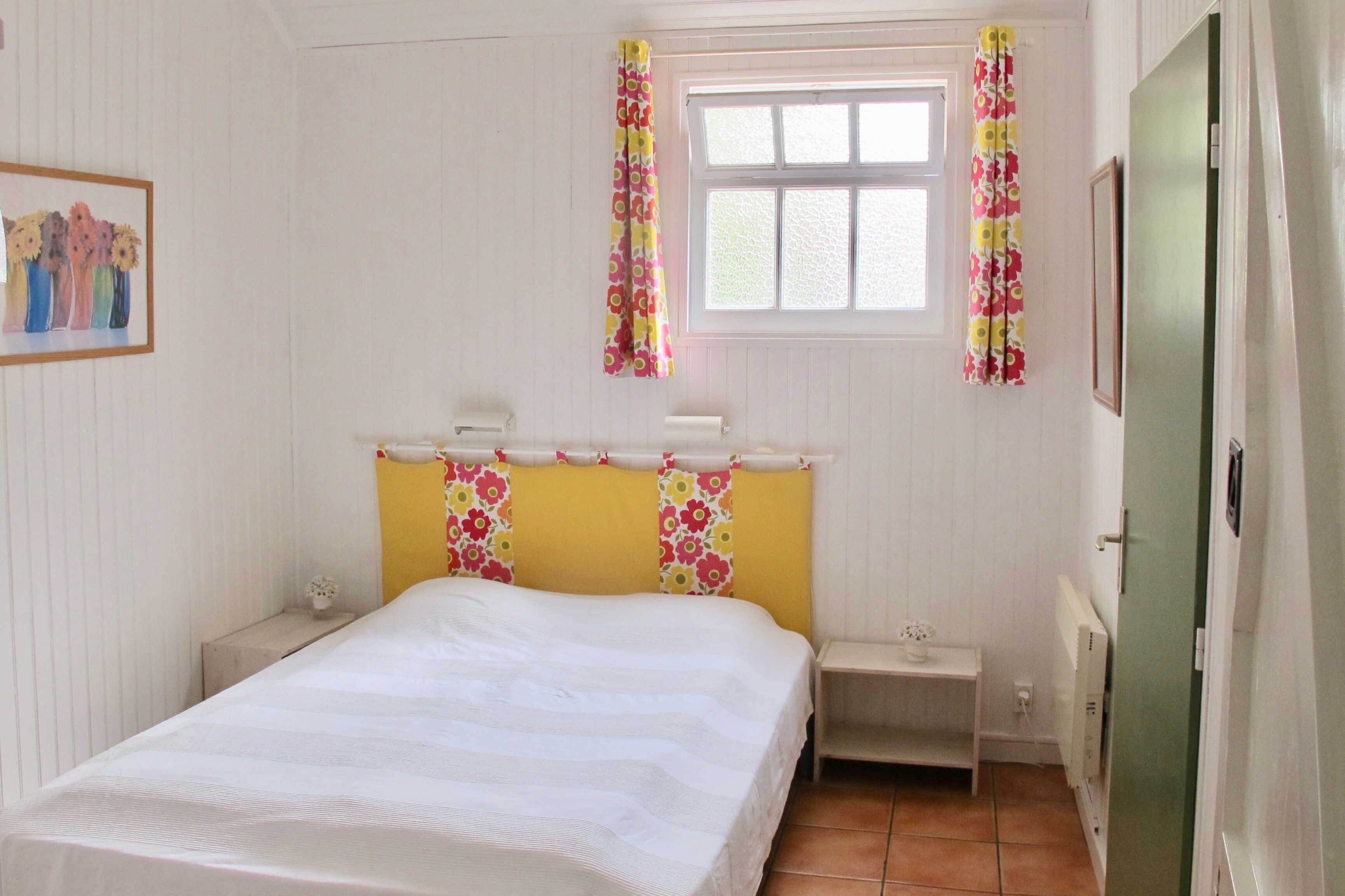Un dormitorio con cama, mesita de noche, ventana con cortinas, radiador y un cuadro en la pared.