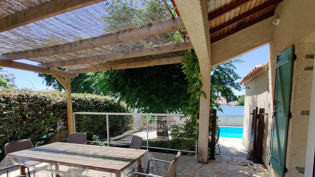 Un patio coperto con tavolo e sedie, una piscina sullo sfondo e vista sul verde circostante.