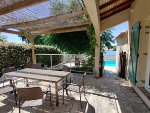 A covered patio with a table and chairs, a pool in the background, and a view of the surrounding greenery. - Pretty single storey villa with private and secure swimming pool 10 km from the beaches (Pomérols)