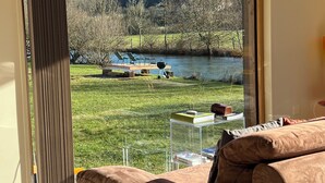 A room with a large sliding glass door leading to a lakeside view, a sofa, and a small table with books. - The Cube is a very private spot for an unforgettable experience by the riverside (Frénois)